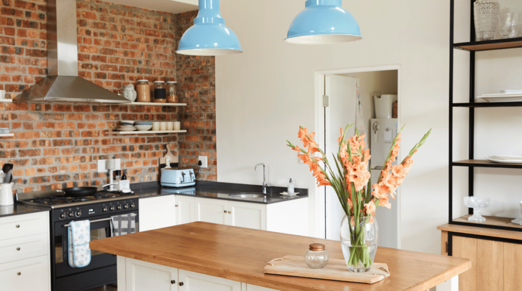 Vue d’une cuisine au style rustique et moderne avec mur en briques, îlot central en bois et luminaires bleus suspendus